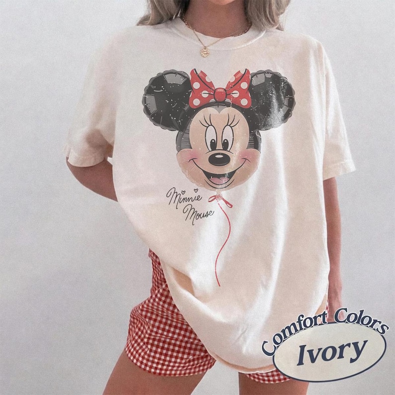 Op de afbeelding: Ivoor kleurig T-shirt met een Minnie Mouse ballon afbeelding, een rode strik en de tekst "Minnie Mouse". Het shirt is van het merk Comfort Colors.