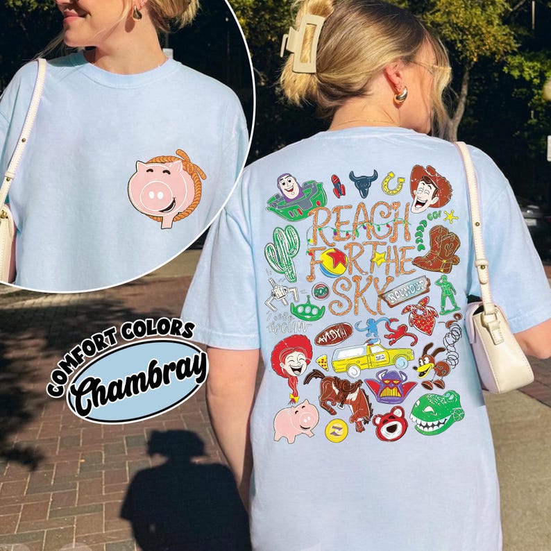 K&ouml;nnte beinhalten: Ein hellblaues T-Shirt mit einer kleinen Schweinegrafik auf der linken Brust. Die R&uuml;ckseite zeigt eine gro&szlig;e Collage aus bunten Zeichentrickfiguren und den Text "REACH FOR THE SKY" in einer stilisierten, strukturierten braunen Schrift.