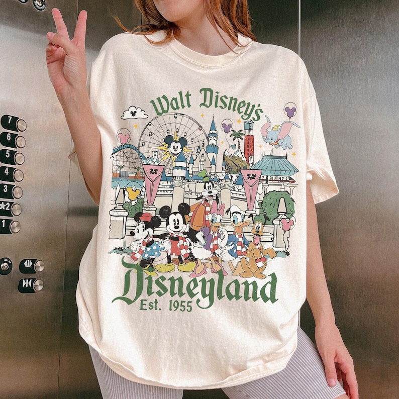 May include: Cream-colored t-shirt featuring a Disneyland graphic with Mickey Mouse, Minnie Mouse, Donald Duck, and other Disney characters. The design includes a Ferris wheel, castle, and the text "Walt Disney's Disneyland Est. 1955."