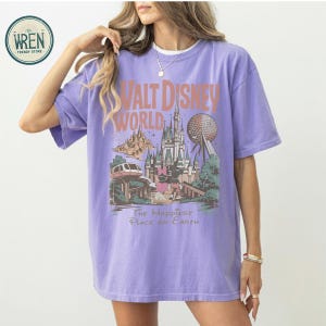 Może przedstawiać: Liliowa koszulka z grafiką w stylu vintage z Walt Disney World. Projekt zawiera zamek, Spaceship Earth oraz tekst "Walt Disney World" i "The Happiest Place on Earth."