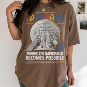May include: A brown short-sleeve t-shirt featuring a graphic of a geodesic dome structure. The text reads "EXPERIMENTAL PROTOTYPE COMMUNITY OF TOMORROW", "SPARK YOUR IMAGINATION", and "WHERE THE IMPOSSIBLE BECOMES POSSIBLE" with small colorful geometric accents.