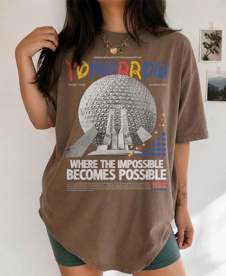 May include: A brown short-sleeve t-shirt featuring a graphic of a geodesic dome structure. The text reads "EXPERIMENTAL PROTOTYPE COMMUNITY OF TOMORROW", "SPARK YOUR IMAGINATION", and "WHERE THE IMPOSSIBLE BECOMES POSSIBLE" with small colorful geometric accents.
