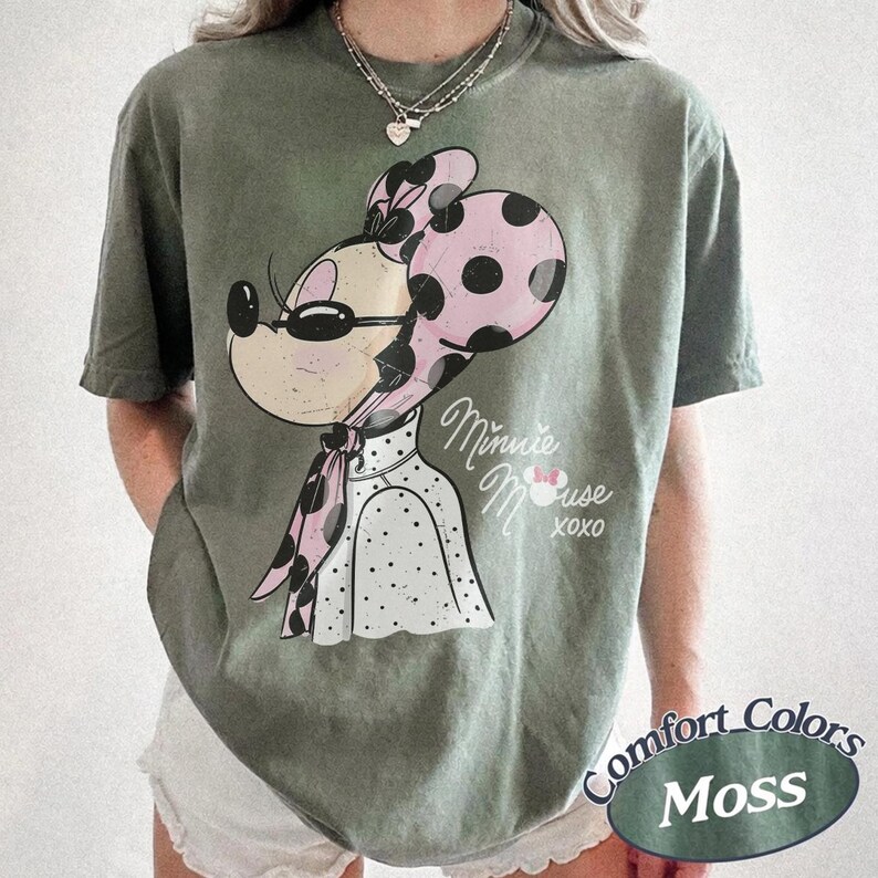May include: Moss green t-shirt featuring a Minnie Mouse graphic. The cartoon character wears sunglasses, a pink polka-dot bow, and a matching scarf. The text "Minnie Mouse XOXO" is printed below the graphic. The shirt has a Comfort Colors label.