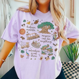 May include: A light purple t-shirt featuring various cartoon illustrations and text. The shirt includes images of animals, vehicles, and the text "hakuna matata", "anyone can be anything", and "adventure is out there".