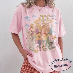 May include: A light pink short-sleeve t-shirt featuring a large graphic print of multiple animated characters in various outfits, including a cowboy hat and space suit. The text TS is printed at the top, with clouds and a rocket ship surrounding the figures.
