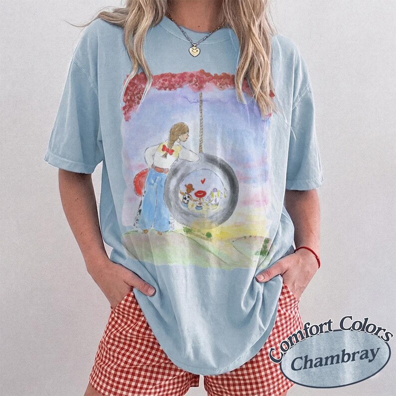 May include: A light blue short-sleeve t-shirt featuring a watercolor-style graphic of a person standing next to a tire swing. The graphic includes a red tree canopy, a blue sky, and a small scene inside the tire. The shirt has a crew neckline.