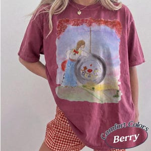 May include: A berry-colored short-sleeve t-shirt featuring a watercolor-style graphic of a person standing next to a large tire swing. Inside the tire, smaller figures are visible. The shirt has a crew neckline and a relaxed, loose-fitting silhouette.