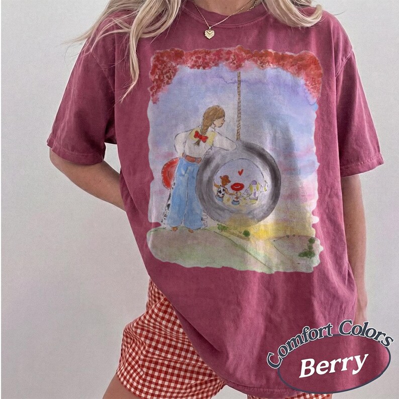 May include: A berry-colored short-sleeve t-shirt featuring a watercolor-style graphic of a person standing next to a large tire swing. Inside the tire, smaller figures are visible. The shirt has a crew neckline and a relaxed, loose-fitting silhouette.