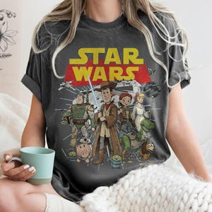 May include: Dark gray t-shirt featuring a Star Wars graphic with characters from Toy Story. The design includes the Star Wars logo in yellow and red, with illustrations of Woody, Buzz Lightyear, and other characters. The shirt has a vintage, distressed look.