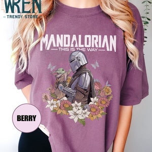 May include: A purple t-shirt featuring a floral design surrounding a graphic of the Mandalorian and Grogu. The text on the shirt reads "Mandalorian - This is the Way - "