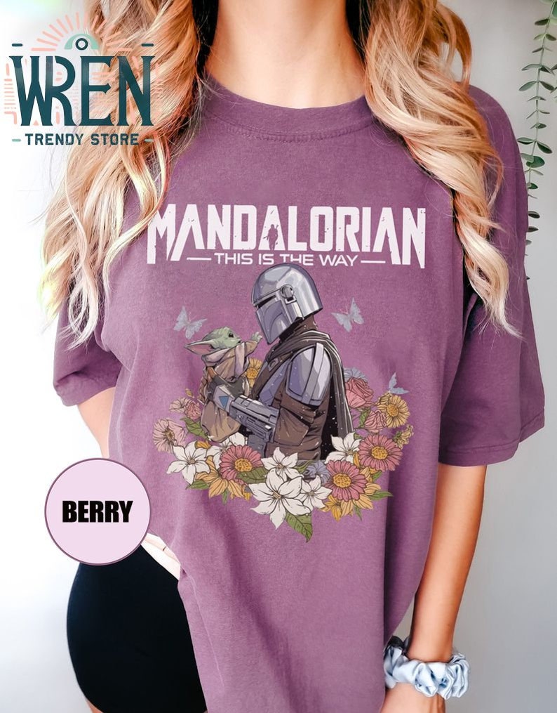May include: A purple t-shirt featuring a floral design surrounding a graphic of the Mandalorian and Grogu. The text on the shirt reads "Mandalorian - This is the Way - "