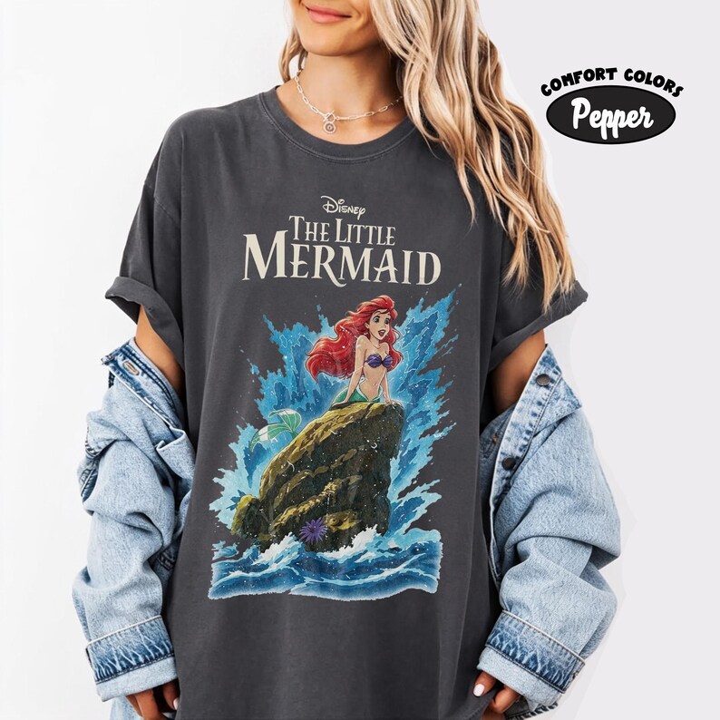 May include: A charcoal gray t-shirt featuring a graphic of Ariel from Disney's The Little Mermaid sitting on a rock amidst blue ocean waves. The text reads Disney The Little Mermaid. A small circular badge in the corner says Comfort Colors Pepper.