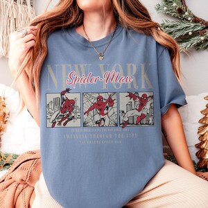 Pu&ograve; includere: Una maglietta blu con una grafica di Spider-Man. Il design include le parole "New York" e "Spider-Man" in un font stilizzato, insieme a pannelli di fumetti di Spider-Man in azione. Il testo aggiuntivo recita "Swinging Through The City."