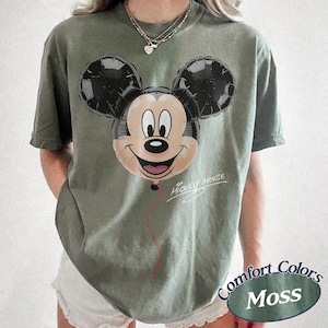 Op de afbeelding: Olijfgroen T-shirt met een Mickey Mouse ballonafbeelding. De ballon heeft zwarte oren en een lachend gezicht. De woorden "Mickey Mouse" zijn cursief geschreven. Het shirt heeft ook een "Comfort Colors Moss" label.