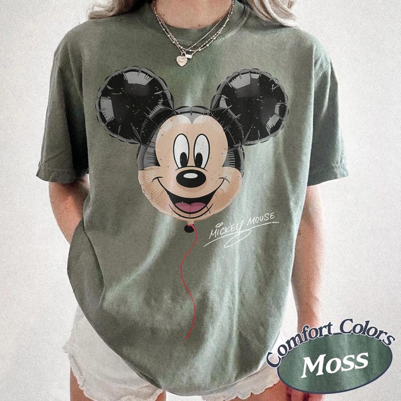 Op de afbeelding: Olijfgroen T-shirt met een Mickey Mouse ballonafbeelding. De ballon heeft zwarte oren en een lachend gezicht. De woorden "Mickey Mouse" zijn cursief geschreven. Het shirt heeft ook een "Comfort Colors Moss" label.