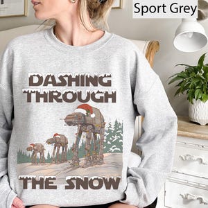 May include: Sport grey sweatshirt with the text "Dashing Through The Snow". The graphic features AT-AT walkers wearing Santa hats, set against a snowy landscape with pine trees. A festive holiday design.