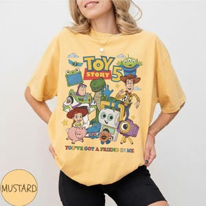 May include: Mustard yellow t-shirt featuring a graphic of characters from Toy Story 5. The design includes Woody, Buzz Lightyear, and other characters. The text "YOU'VE GOT A FRIEND IN ME" is printed below the characters.
