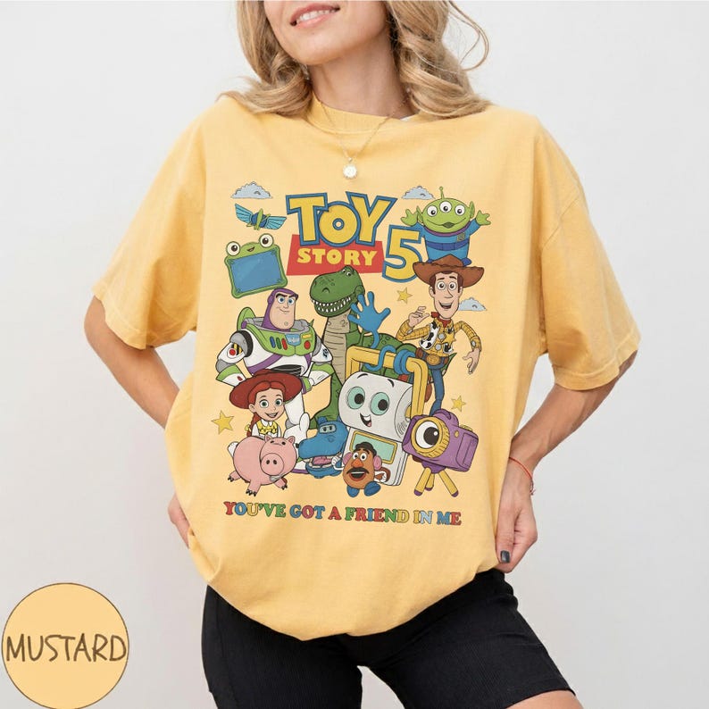 May include: Mustard yellow t-shirt featuring a graphic of characters from Toy Story 5. The design includes Woody, Buzz Lightyear, and other characters. The text "YOU'VE GOT A FRIEND IN ME" is printed below the characters.