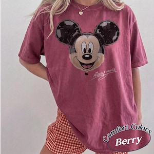 Op de afbeelding: Bessenkleurig T-shirt met een Mickey Mouse ballonafbeelding. Het ontwerp bevat de woorden "Mickey Mouse" in script. Het shirt is gelabeld met "Comfort Colors Berry" in een afgeronde ovaal.