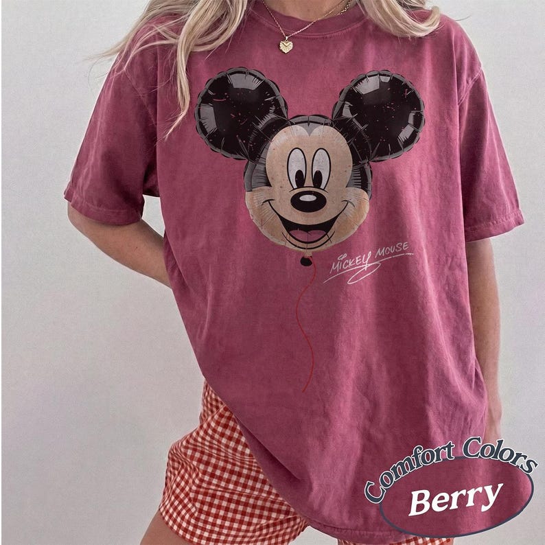 Op de afbeelding: Bessenkleurig T-shirt met een Mickey Mouse ballonafbeelding. Het ontwerp bevat de woorden "Mickey Mouse" in script. Het shirt is gelabeld met "Comfort Colors Berry" in een afgeronde ovaal.