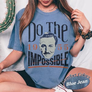 May include: Blue jean-colored t-shirt with the text "Do The Impossible" and a portrait of a man. The numbers "1955" are printed above the word "Impossible". The shirt is a Comfort Colors brand.