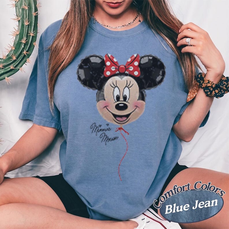 Op de afbeelding: Blauw Comfort Colors T-shirt met een Minnie Mouse ballon ontwerp. De afbeelding toont Minnie Mouse met een rode strik en de woorden "Minnie Mouse". Het shirt is blauw van kleur.