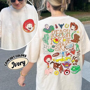 K&ouml;nnte beinhalten: Ein elfenbeinfarbenes T-Shirt mit einer kleinen Grafik einer rothaarigen Figur auf der linken Brust. Die R&uuml;ckseite zeigt eine gro&szlig;e, bunte Collage aus verschiedenen Zeichentrickfiguren und Objekten mit dem Text REACH FOR THE SKY in gro&szlig;en Buchstaben.