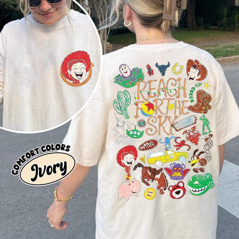 K&ouml;nnte beinhalten: Ein elfenbeinfarbenes T-Shirt mit einer kleinen Grafik einer rothaarigen Figur auf der linken Brust. Die R&uuml;ckseite zeigt eine gro&szlig;e, bunte Collage aus verschiedenen Zeichentrickfiguren und Objekten mit dem Text REACH FOR THE SKY in gro&szlig;en Buchstaben.