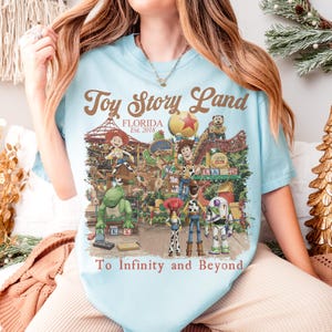 May include: Light blue t-shirt featuring a colorful graphic of Toy Story Land in Florida. The design includes various Toy Story characters and the text "Toy Story Land Florida Est. 2018" and "To Infinity and Beyond."