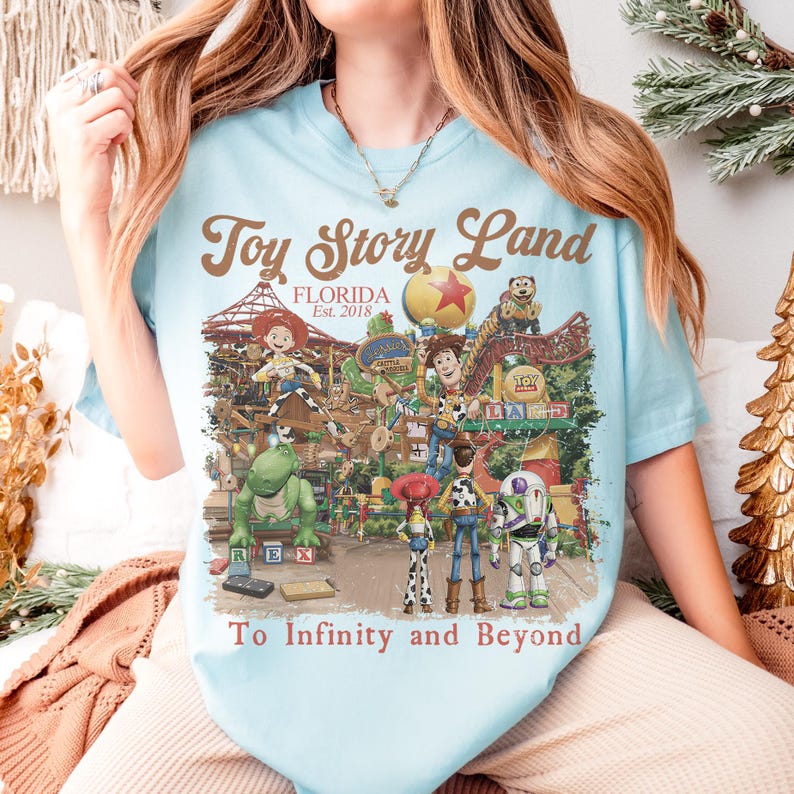May include: Light blue t-shirt featuring a colorful graphic of Toy Story Land in Florida. The design includes various Toy Story characters and the text "Toy Story Land Florida Est. 2018" and "To Infinity and Beyond."