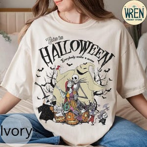 以下が含まれることがあります： ハロウィーンをテーマにしたアイボリー色のTシャツ。デザインには「This is Halloween」と「Everybody make a scene」の文字が含まれています。グラフィックには、ジャック・スケリントン、サリー、ウーギー・ブギーなどのキャラクター、コウモリ、カボチャ、魔女などが描かれています。