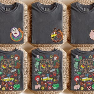 K&ouml;nnte beinhalten: Ein anthrazitfarbenes T-Shirt mit einer kleinen Charaktergrafik auf der linken Brust und einer gro&szlig;en, bunten Collage aus Toy Story-Figuren und Symbolen auf der R&uuml;ckseite, inklusive des Textes "REACH FOR THE SKY" in Western-Schrift.
