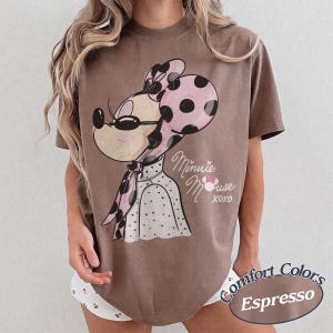 May include: Espresso-colored t-shirt featuring a vintage-style Minnie Mouse graphic. Minnie is depicted wearing a pink polka-dot bow and scarf, with the words "Minnie Mouse XOXO" printed below. The shirt is a Comfort Colors brand.