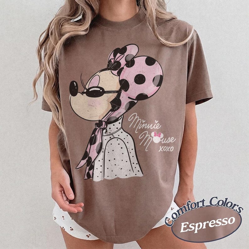 May include: Espresso-colored t-shirt featuring a vintage-style Minnie Mouse graphic. Minnie is depicted wearing a pink polka-dot bow and scarf, with the words "Minnie Mouse XOXO" printed below. The shirt is a Comfort Colors brand.