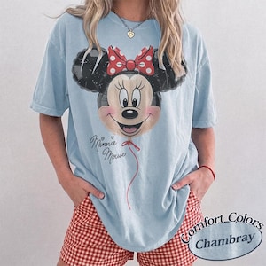 Op de afbeelding: Lichtblauw T-shirt met een Minnie Mouse ballonafbeelding, een rode strik en de tekst "Minnie Mouse". De outfit bevat rood-witte geruite shorts. Het shirt is van Comfort Colors.