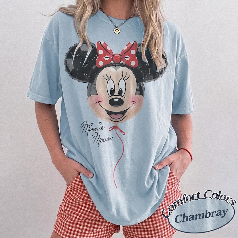 Op de afbeelding: Lichtblauw T-shirt met een Minnie Mouse ballonafbeelding, een rode strik en de tekst "Minnie Mouse". De outfit bevat rood-witte geruite shorts. Het shirt is van Comfort Colors.