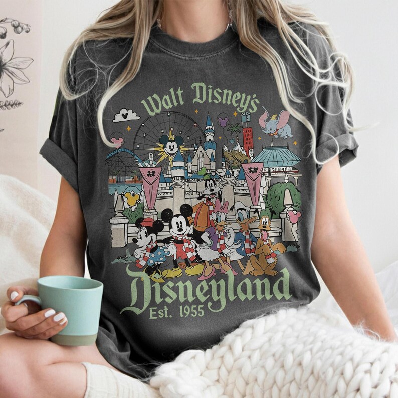 May include: Dark gray t-shirt featuring a vintage Disneyland design. The graphic includes Mickey and Minnie Mouse, Donald Duck, and other Disney characters in front of park landmarks. The text "Walt Disney's Disneyland Est. 1955" is also displayed.