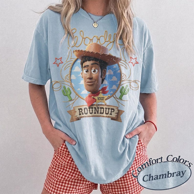 K&ouml;nnte beinhalten: Ein hellblaues Kurzarm-T-Shirt mit einer Grafik der Figur Woody aus Toy Story in einem Seilkreis. Das Design enth&auml;lt den Text Woody, Roundup und einen Sheriffstern sowie kleine Illustrationen von Kakteen und Sternen.