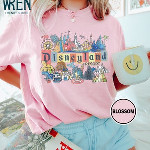 May include: Pink t-shirt with a colorful graphic of the Disneyland Resort logo and various attractions. The graphic includes the text "Disneyland Resort".