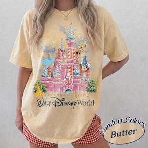 Może przedstawiać: Koszulka w kolorze maślanym z kolorową grafiką zamku Kopciuszka i postaciami Disneya. Tekst "This is Now! Walt Disney World 1997" jest wydrukowany poniżej. Marka Comfort Colors.