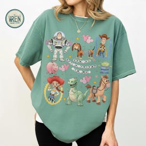 May include: Sage green t-shirt with cartoon characters from a popular animated film. The design includes Buzz Lightyear, Woody, Jessie, and other characters. The text on the shirt reads "YOU'VE GOT A FRIEND IN ME."