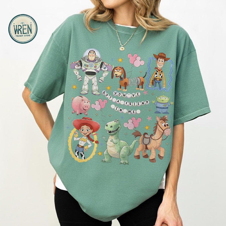 May include: Sage green t-shirt with cartoon characters from a popular animated film. The design includes Buzz Lightyear, Woody, Jessie, and other characters. The text on the shirt reads "YOU'VE GOT A FRIEND IN ME."