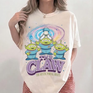 以下が含まれることがあります： クリーム色のTシャツ。青い衣装を着た3人の緑色のエイリアンのグラフィックと、紫色の「THE CLAW」の文字が特徴です。背景にはカラフルな銀河のデザインがあり、その下には「THE MYSTIC PORTAL AWAITS」の文字があります。