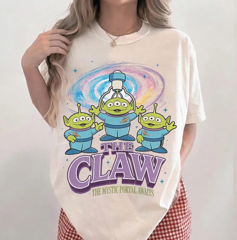 以下が含まれることがあります： クリーム色のTシャツ。青い衣装を着た3人の緑色のエイリアンのグラフィックと、紫色の「THE CLAW」の文字が特徴です。背景にはカラフルな銀河のデザインがあり、その下には「THE MYSTIC PORTAL AWAITS」の文字があります。