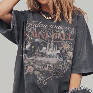 Puede incluir: Una camiseta gris carb&oacute;n de corte oversize con un estampado gr&aacute;fico de un castillo, flores y mariposas. El texto dice Today was a FAIRY TALE y Can you feel this magic in the air? Un peque&ntilde;o logotipo circular indica Comfort Colors Pepper.