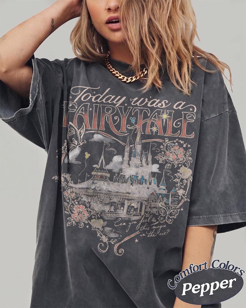 Puede incluir: Una camiseta gris carb&oacute;n de corte oversize con un estampado gr&aacute;fico de un castillo, flores y mariposas. El texto dice Today was a FAIRY TALE y Can you feel this magic in the air? Un peque&ntilde;o logotipo circular indica Comfort Colors Pepper.