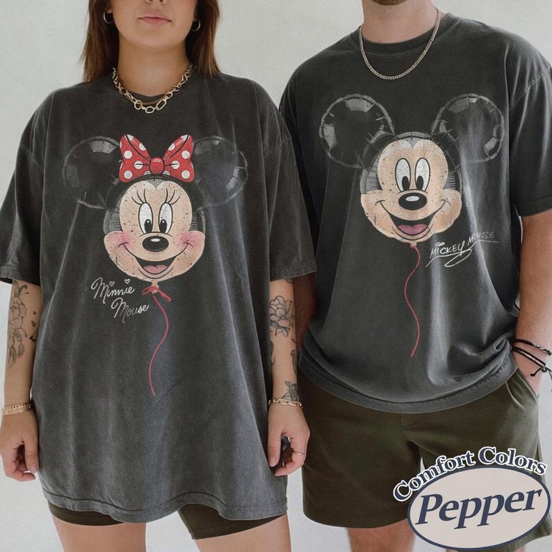 Op de afbeelding: Twee antracietgrijze t-shirts met Disney-personages. E&eacute;n shirt toont Minnie Mouse met een rode strik met stippen, de andere Mickey Mouse. Beide ontwerpen zijn gedrukt als ballonnen. De shirts zijn gelabeld met "Comfort Colors Pepper".