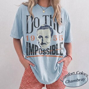 May include: Light blue t-shirt with the text "DO THE IMPOSSIBLE" and a vintage image of a man's face. The shirt also features the numbers "1955". The shirt is part of the Comfort Colors Chambray collection.