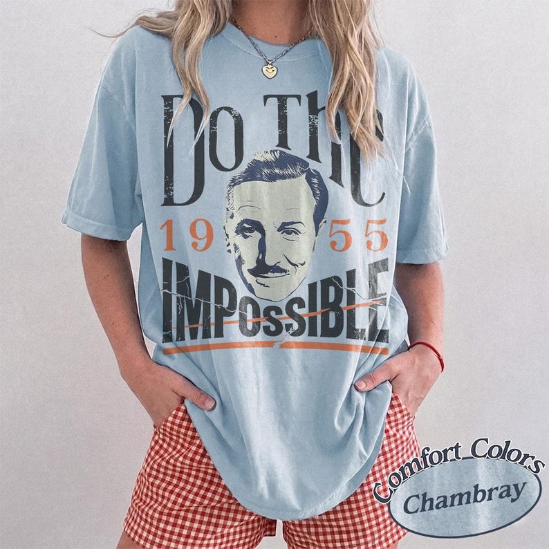 May include: Light blue t-shirt with the text "DO THE IMPOSSIBLE" and a vintage image of a man's face. The shirt also features the numbers "1955". The shirt is part of the Comfort Colors Chambray collection.