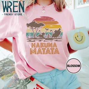 May include: A pink t-shirt with a vintage sunset graphic featuring the Lion King characters Simba, Timon, and Pumbaa. The text "Hakuna Matata" is printed below the graphic.